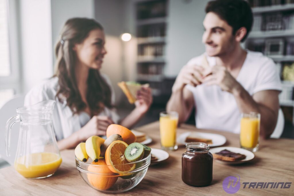 Young romantic couple is having breakfast on modern light kitchen. Sexy woman in underwear and men's shirt and handsome man are sitting at the table eating sandwiches and toasts drinking orange juice. Healthy lifestyle concept.